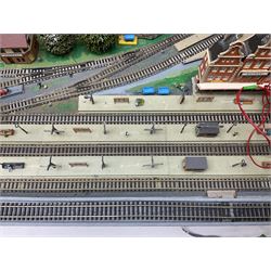 Table top 'N' gauge continental scenic layout as a split level town with various loops of track, station with numerous platforms, engine shed, assorted buildings, roads with motor vehicles and figures, tunnels, trees, river with bridge, tram depot etc 122 x 52cm