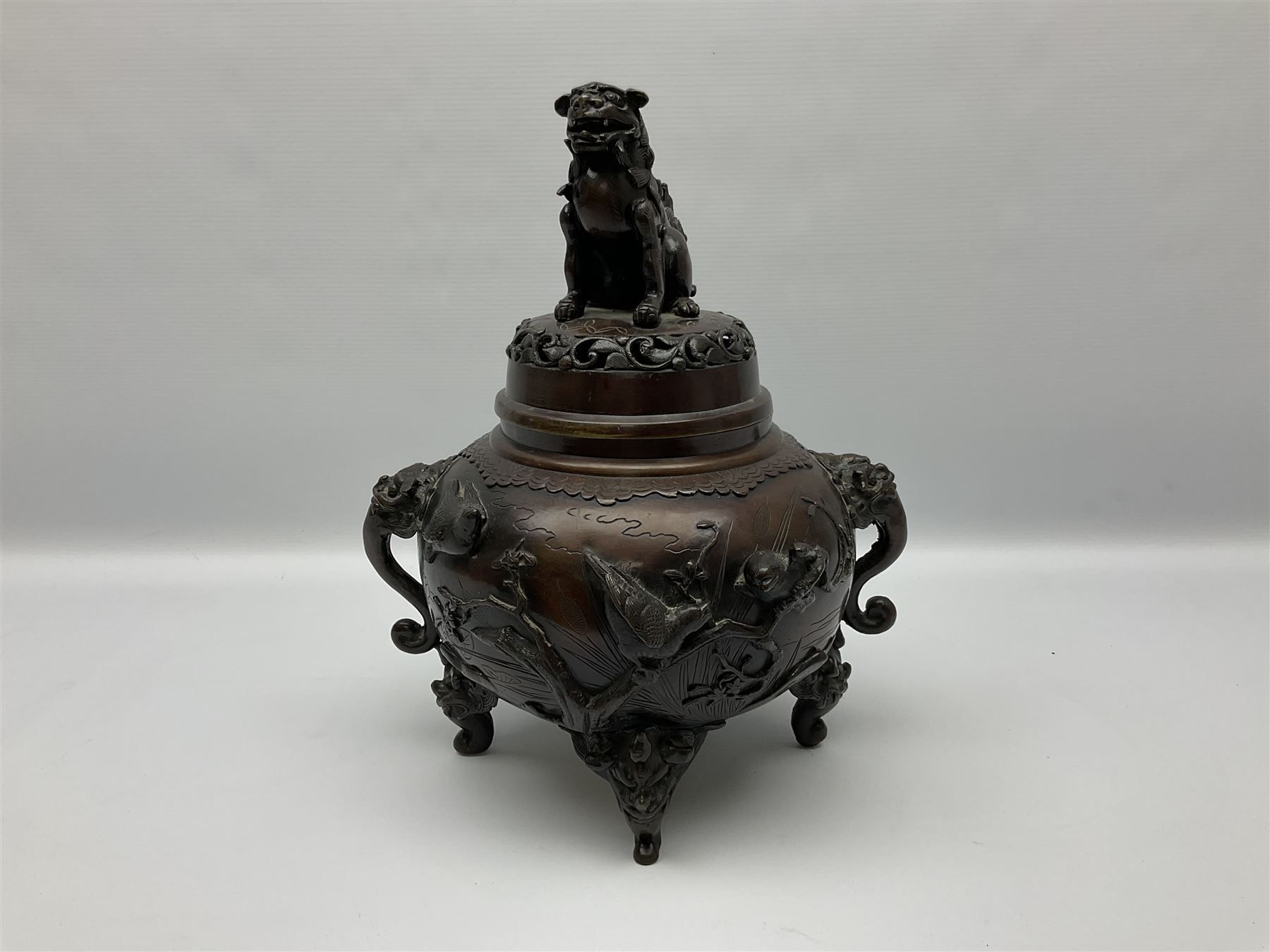 Japanese bronzed koro, the body of globular form with twin cast zoomorphic mask handles, decorated in relief with birds upon blossoming branches, against a ground incised with rushes and peonies, the flat toped domed cover with pierced band and surmounted by a cast figure of a Foo dog, the whole upon three zoomorphic mask feet, with impressed character type mark beneath, H35.5cm