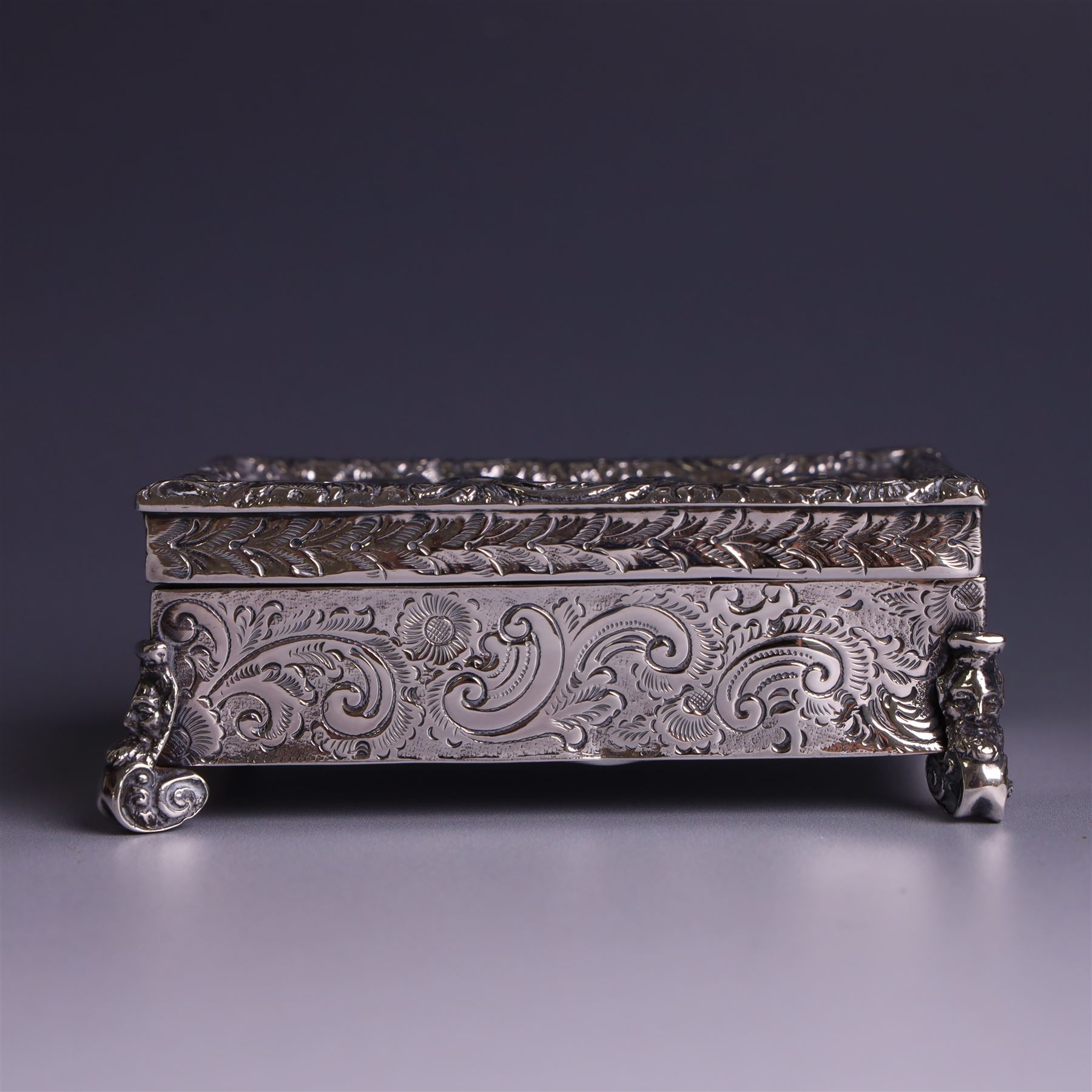 Victorian silver tobacco box, rectangular form, hinged lid embossed with a gaming scene, the body engraved with scrolls and flowers, upon four scrolling mask feet, hallmarked George Heath London 1885, H4.5cm, W10.5cm