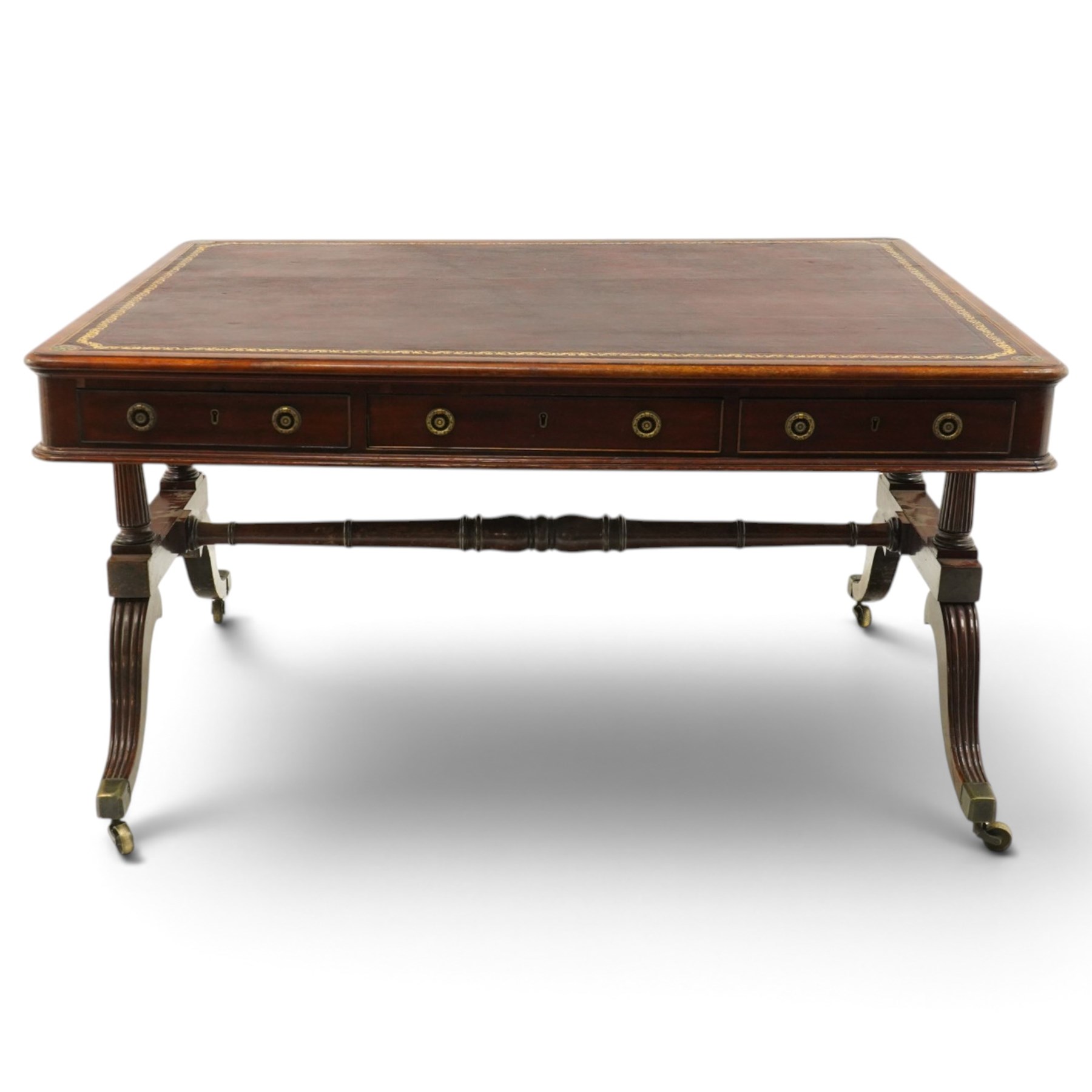 Regency inlaid mahogany and brass mounted library table, the rectangular top inset with gilt tooled burgundy leather writing surface, enclosed by  crossbanding and boxwood stringing with roundel corner motifs, above three cock-beaded frieze drawers to either side fitted with brass ring handles and brass escutcheons, the end friezes centred by applied brass foliate mounts, raised on twin reeded column supports over rectangular platform ends, united by a ring-turned stretcher, the reeded sabre legs terminating in brass caps and castors, one drawer bearing ivorine label 'Spillman & Co., St Martins Lane, London'