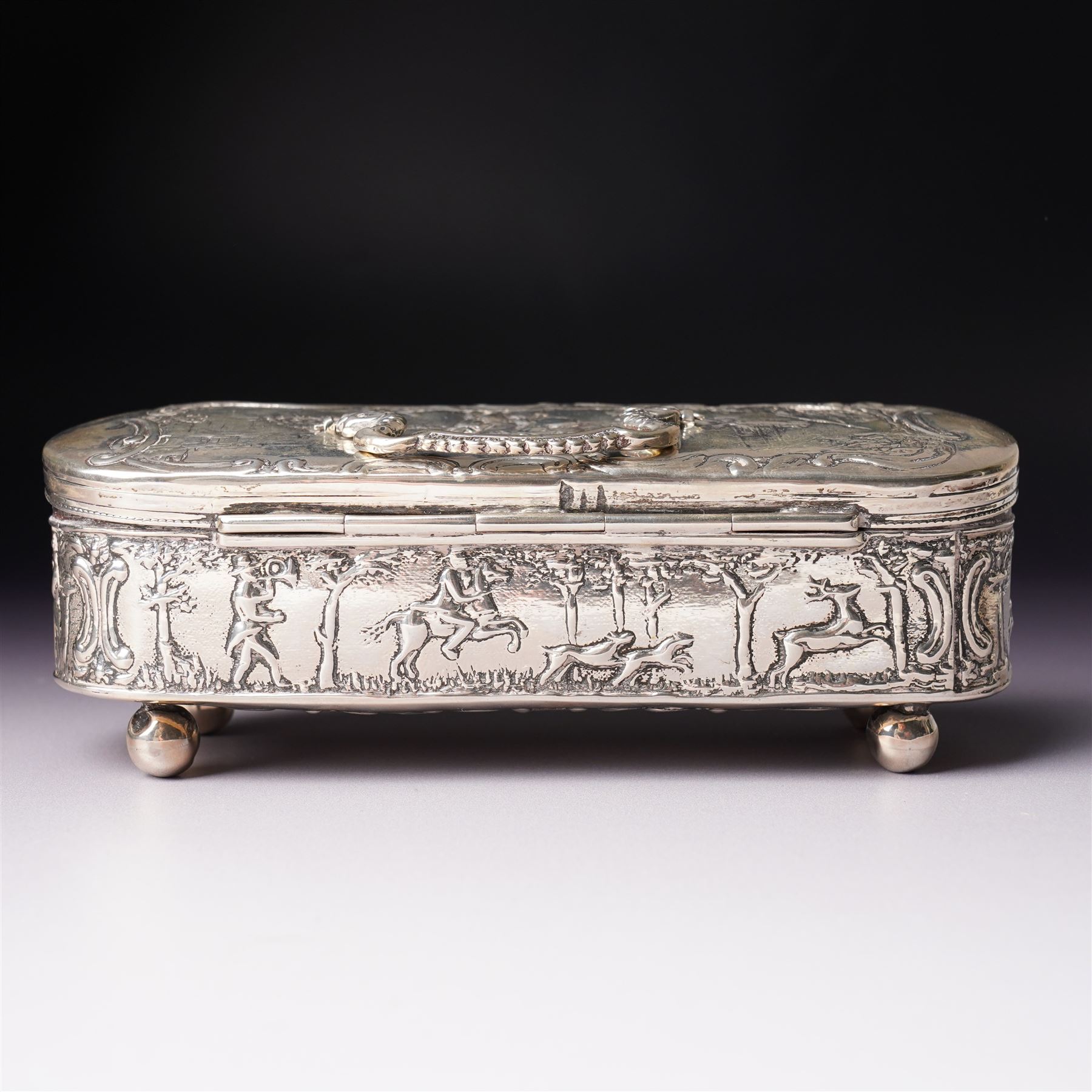19th century continental silver box of oval design , the hinged cover with swing handle embossed with sailing ships, castle etc, the base decorated with horse and cart in rural landscape and the sides with a hunting scene on ball feet L15cm
