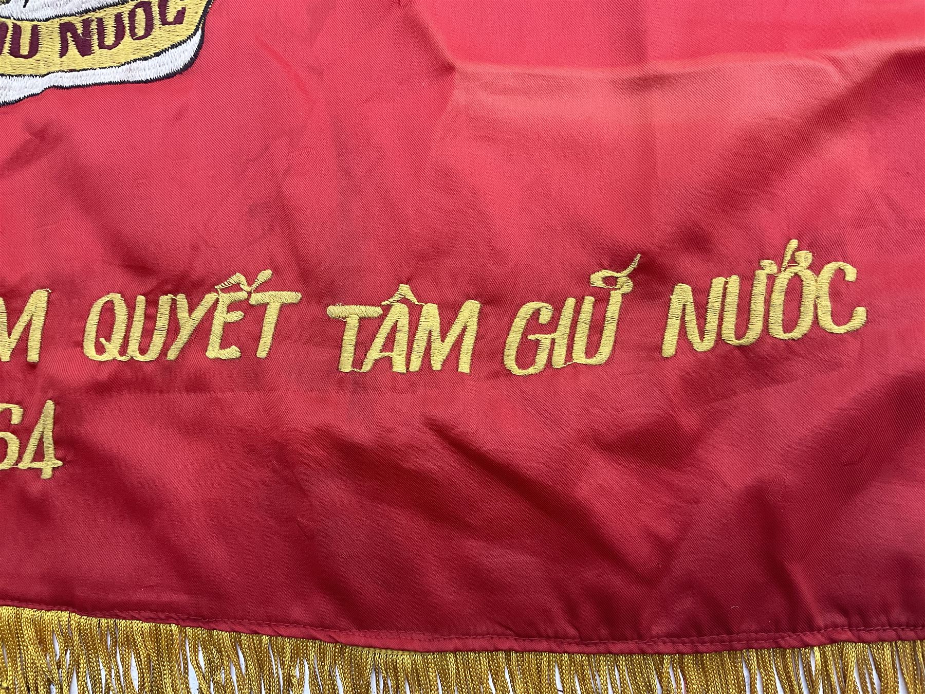 1960s North Vietnam banner embroidered in yellow thread on a red ground, roughly translates as 'Peoples Army of Viet Nam. Warriors of Viet Nam Determined To Keep The Country 1964' around a central military crest with the motto 'Warriors Keep The Country'; tassels to three sides 70 x 100cm