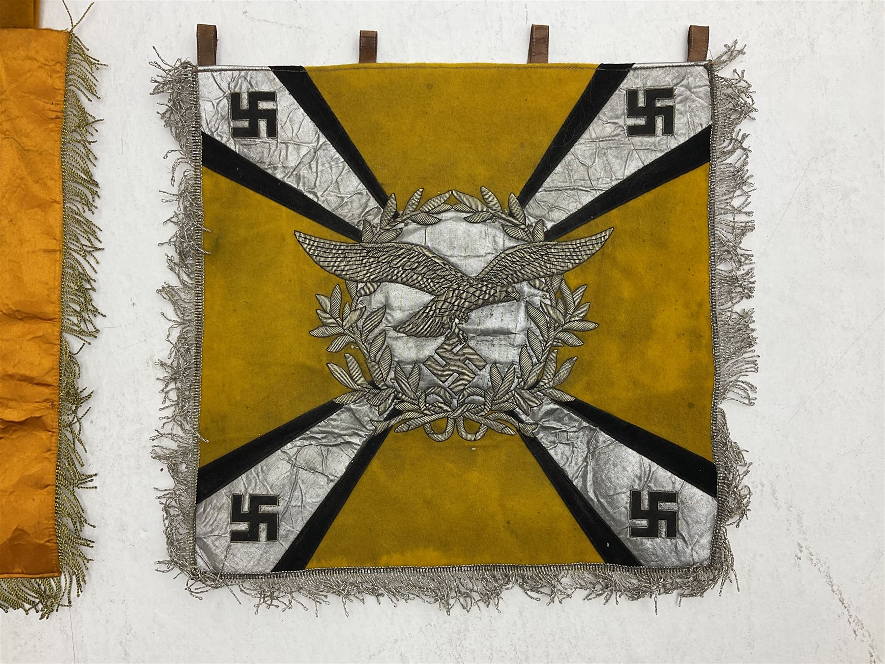 Two replica German Luftwaffe trumpet banners, one heavily silver wire embroidered with Luftwaffe eagle in a wreath, on an amber, white and black rayed background, , with silver wire tasselled border and four leather hanging loops, approx. 50 x 49cm; the other slightly larger heavily gold wire embroidered with the Luftwaffe eagle on a plain amber ground with gold wire tasselled border and two hanging loops (2)