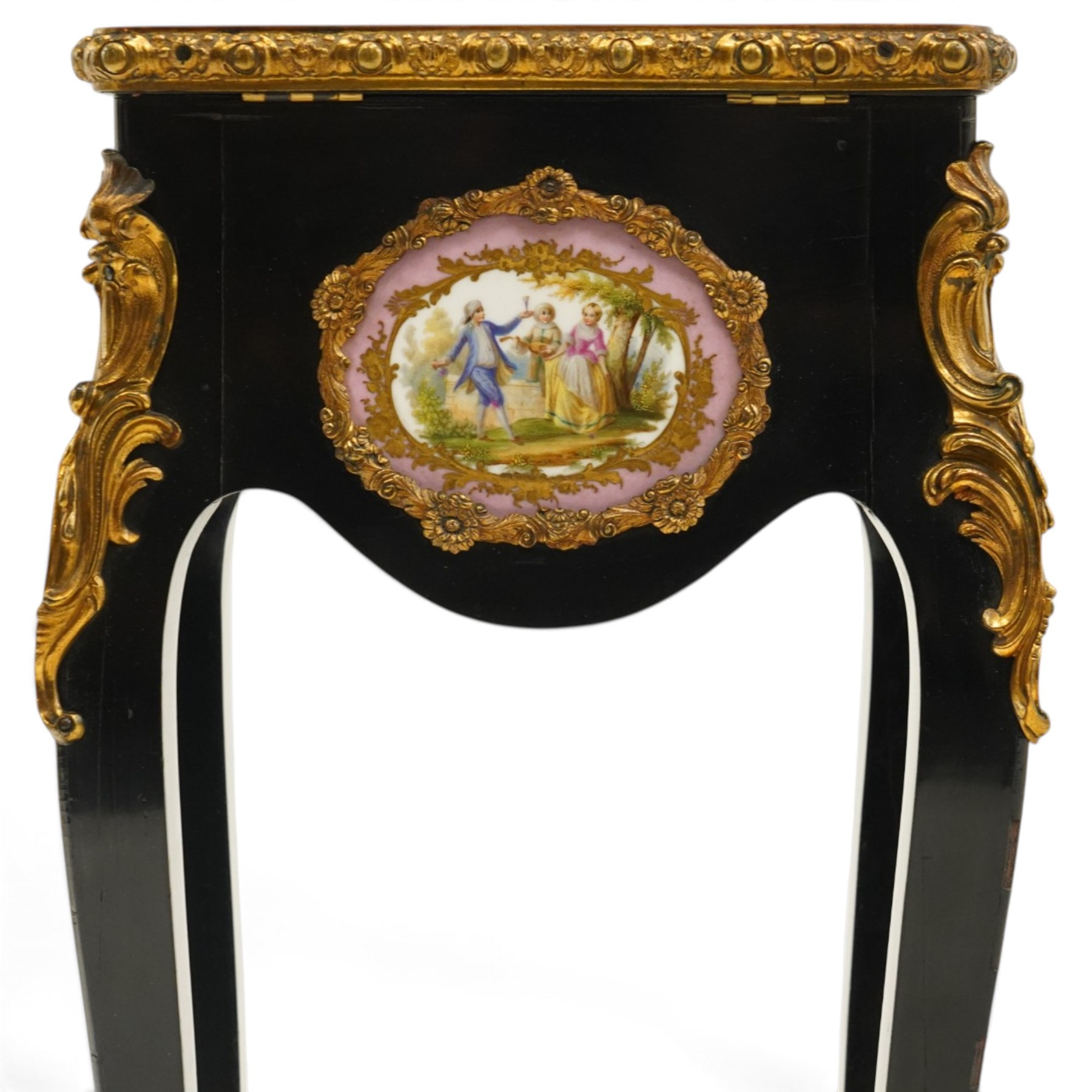 Early 20th century French ebonised sewing or work table, rectangular hinged top with foliate moulded gilt metal edge, enclosing upholstered interior, over a central Sevres style porcelain plaque depicting a courting scene, raised on cabriole supports with gilt metal carotuche and acanthus knee mounts, united by undertier with brass gallery terminating to gilt metal scroll feet