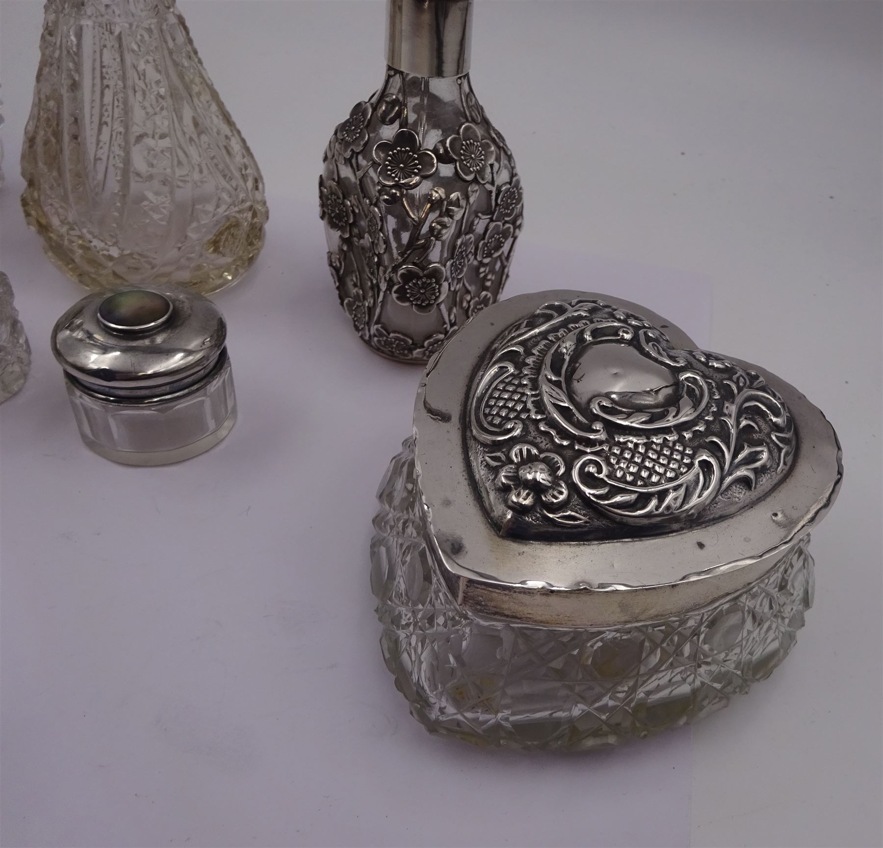 Two silver mounted dressing table jars, one in the form of a heart, both with embossed silver covers and cut glass bodies, hallmarked, together with two smaller examples, one set with mother of pearl, three silver mounted glass scent bottles, including an example with prunus blossom decoration, stamped Sterling, tallest bottle H17cm