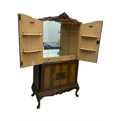 Early 20th century walnut veneered cocktail cabinet, shaped and carved foliate pediment above a pair of arched panelled doors with figured veneers and ornate cast escutcheons, interior is mirrored on the base and back, slide out mirrored shelf, flanked by foliate carved uprights, over a conforming lower cabinet, all raised on shaped apron and scroll carved cabriole feet