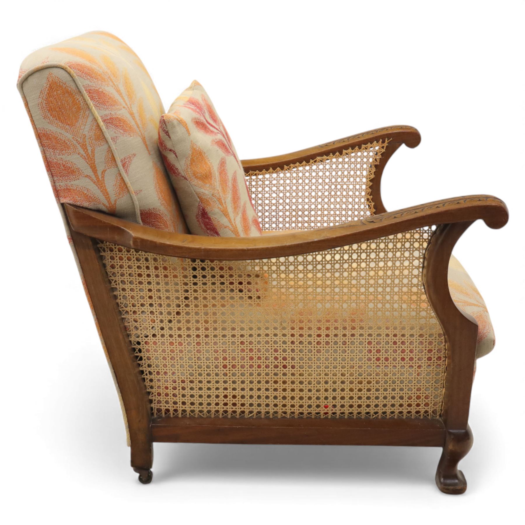 Pair of mid-20th century mahogany framed bergère armchairs, each with carved scroll arms and caned side panels, floral upholstered loose cushions, raised on cabriole front supports