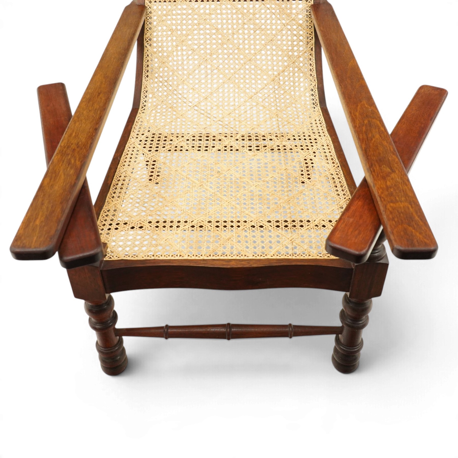 20th century teak framed plantation chair, plain cresting rail over cane back and seat, fitted with hinged extending leg rests, raised on turned front-supports united by ring-turned stretcher
