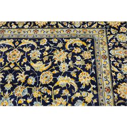 Central Persian Kashan hand-knotted carpet, deep indigo field with a densely all-over arrangement of scrolling floral vines, palmettes, and flowering sprays, in soft ivory, pale blue, and warm gold tones, principal ivory border with a continuous meandering vine of stylized palmettes, and floral motifs, flanked by multiple narrow guard borders, with cotton fringe ends 