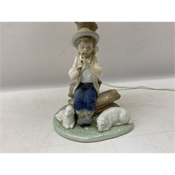 Nao table lamp modelled as a boy playing the flute upon logs with two recumbent sheep at his feet, the stem modelled as a tree trunk, with fabric shade, with printed mark beneath, H46cm