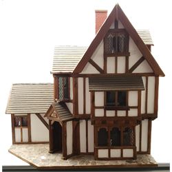 Gerry Welch for Manorcraft Tudor style wooden doll's house, of three-storey multi gabled form with mock beam and plaster walls and simulated leaded glass windows under a felt tiled roof, boarded floors and stairs to each room, curtains to most windows, front entrance door, rear door and single storey extension for kitchen. Furniture includes fitted floral bathroom suite to top floor, well stocked Welsh dresser with copperware etc, tapestry style wall hangings, knole settee, tin bath, metal range, fire-surround, wooden three-piece suite etc; on shaped cobbled base W91cm D61cm H91cm