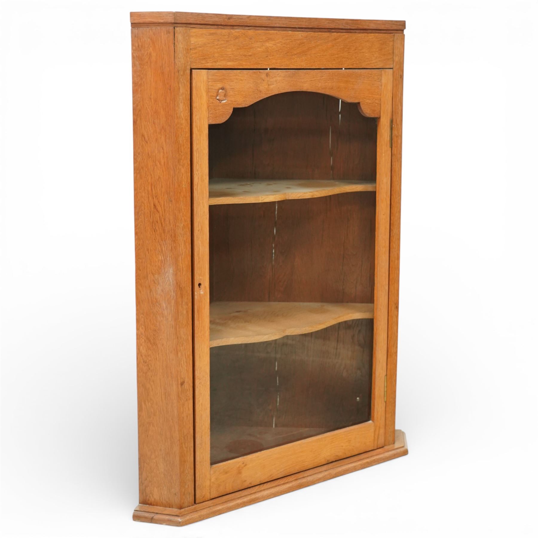 Yorkshire oak corner cabinet, chamfered triangular top above a single glazed door with shaped arched upper rail, enclosing two shaped fixed shelves, plain sides and back, raised on a moulded plinth base, carved with a bell signature