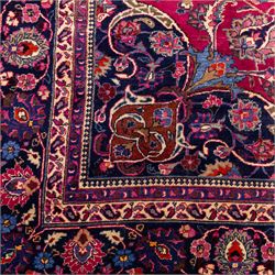 North East Persian Mashad hand-knotted carpet, rich madder red field with central cusped foliate medallion with issuing scrolling arabesques, palmettes, and floral sprays, enclosed within a broad indigo main border of bold palmettes, between multiple guard stripes, with cotton fringe ends 