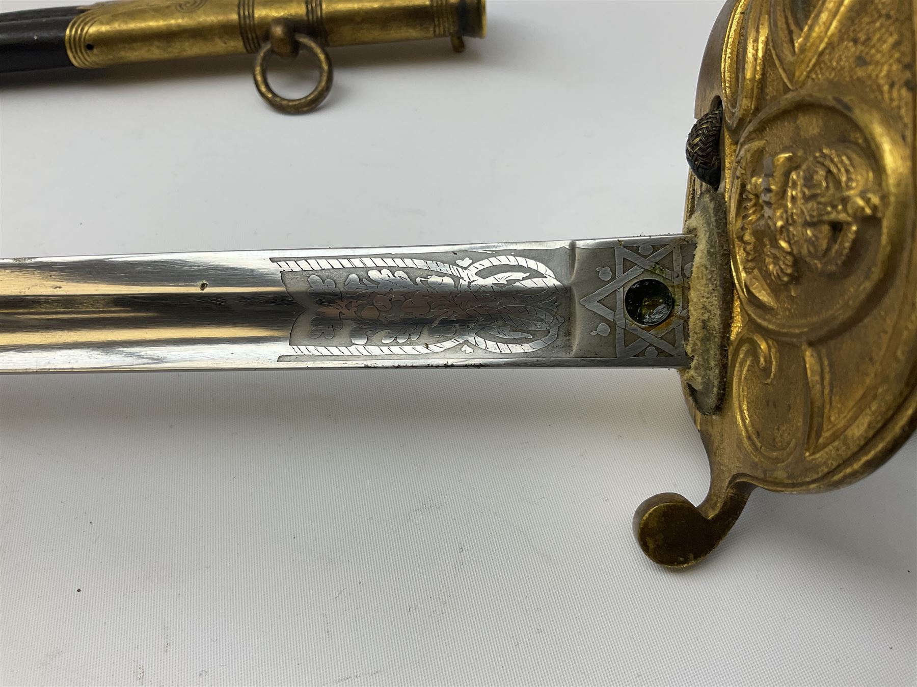 WW1 George V era 1827 Pattern Royal Naval Officer’s sword, the 80cm fullered steel blade decorated with crown, fouled anchor etc and marked Gieves Portsmouth London Edinburgh Devonport to the ricasso, gilded hilt with fouled anchor and crown, wire-bound ray skin grip, lion’s head pommel with mane backstrap, folding/locking langet and full bullion knot; in  leather scabbard having twin brass lockets with hand engraved panels and twin suspension rings L94cm overall. Provenance: By direct family descent from Brian Heathcote HMS Conqueror later Superintendant in the Liverpool Special Constabulary.