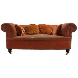 Late Victorian two-seat sofa, traditionally shaped with rolled arms, upholstered in rust fabric with blue and green entwined piping, together with scatter cushions, on turned front feet with brass and ceramic castors