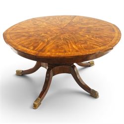 Theodore Alexander - Regency Revival figured mahogany 'Althorp Patent Jupe' extending dining table, the segmented circular top formed of six pull-out triangular divisions with inset fold-out leaves, raised on a large turned vasiform pedestal on a pentagonal platform base with ebonised stringing, terminating to five splayed supports with gilt metal acanthus scrolled feet, retailed by David Hick Interiors, Jersey