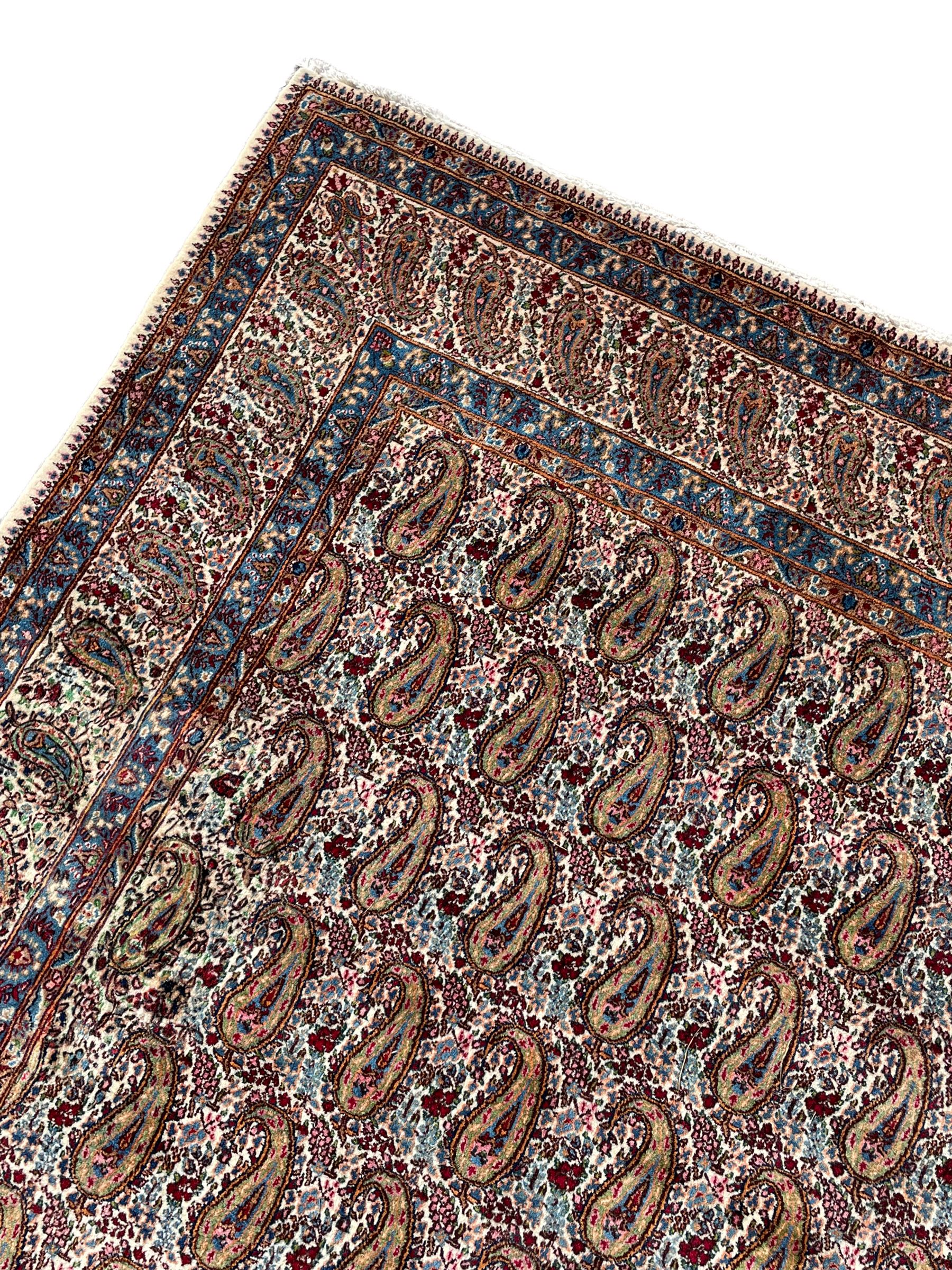 Persian Kirman carpet, the ivory ground field and border decorated with repeating Boteh motifs, interspaced with all-over floral design, blue ground border guards, the top border with signature panel