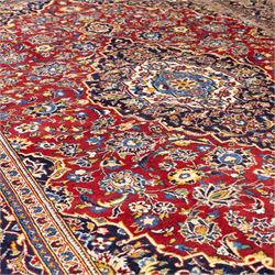 Central Persian Kashan hand-knotted rug, rich red field with a central lobbed medallion surrounded by finely drawn scrolling floral sprays, palmettes, and rosettes, enclosed by a broad dark blue floral border with multiple guard stripes and cotton fringe ends 