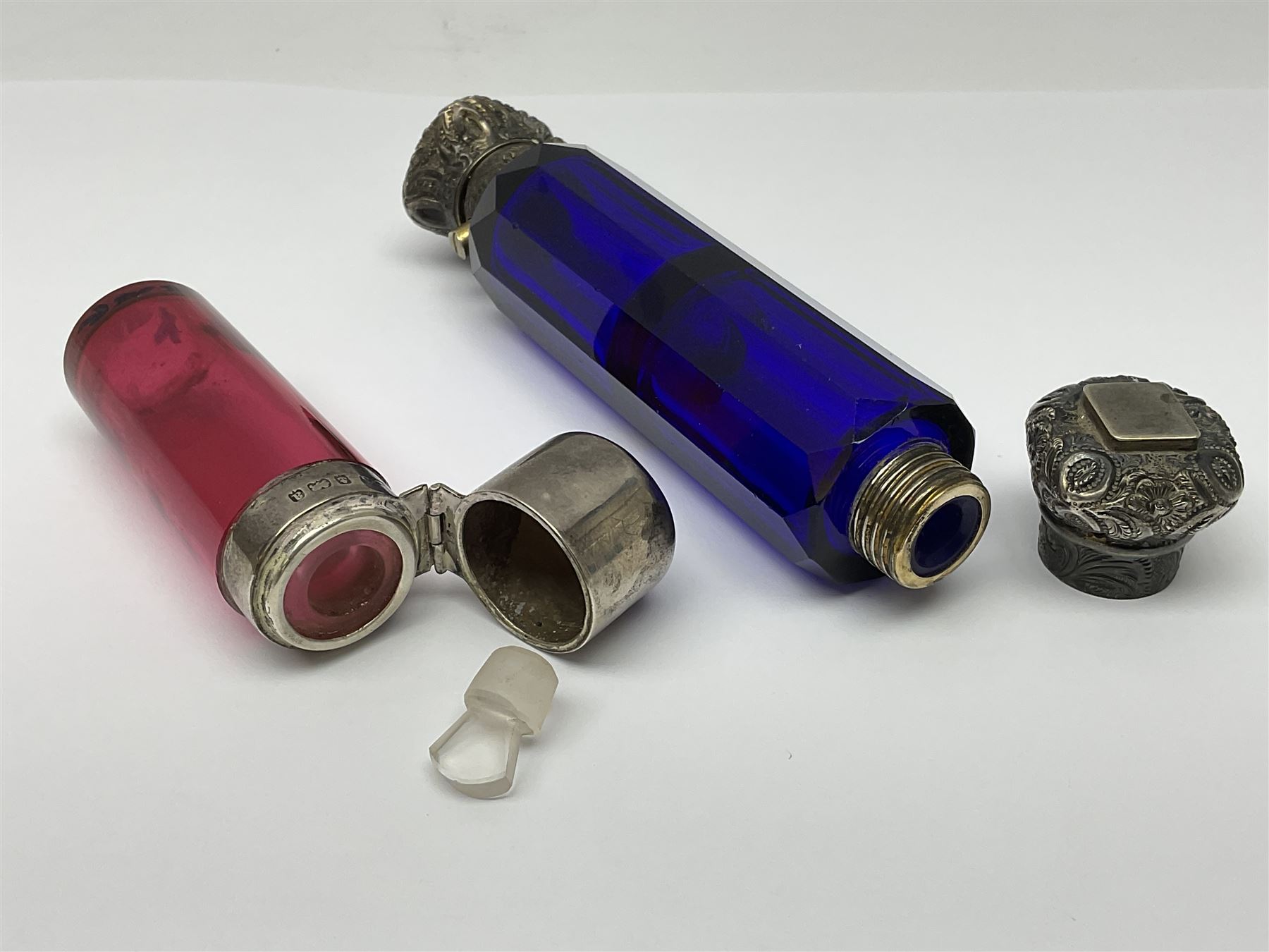 Late Victorian silver mounted ruby glass cylindrical scent bottle, with interior glass stopper, Birmingham 1900, makers mark worn, H8cm, together with a Victorian double ended cut blue glass perfume bottle mounted with white metal screw and flip caps decorated with foliate repousse detailing, H13.5cm