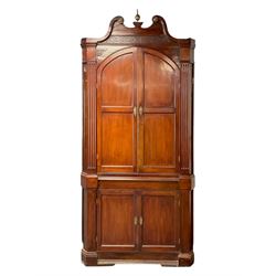 George III mahogany corner cabinet, the scrolled pediment surmounted by a brass finial and carved fretwork frieze, with fluted uprights flanking arched double doors over a painted interior fitted with shaped shelves, above a lower cupboard with twin panelled doors, on plinth base