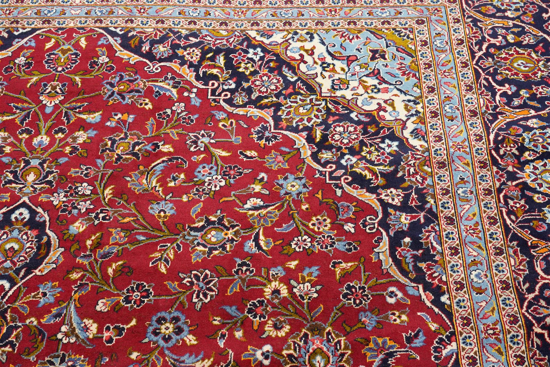 Central Persian Kashan hand-knotted carpet, crimson ground field, with central cusped lozenge medallion, enclosing a palmette and a floral rosette arrangement, surrounding field densely populated with scrolling arabesques, flowering vinery and stylized boteh motifs in tones of indigo, pale blue, ivory and ochre and spandrels of confirming design, flanked by multiple guard stripes, with cotton fringe ends 