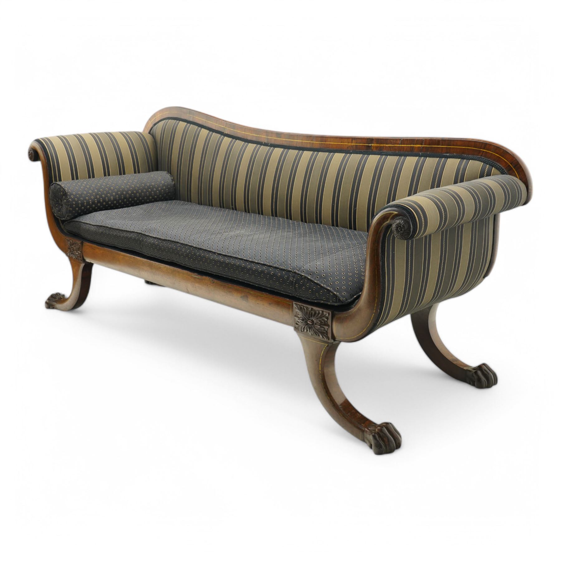 Regency brass inlaid rosewood chaise lounge or settee, shaped back over rolled head and foot, upholstered in black and gold striped fabric with rectangular squab cushion and bolster cushion, the rolled terminals carved with acanthus leaf roundels, lower rectangular foliate carved panels, on out splayed supports with paw carved feet 