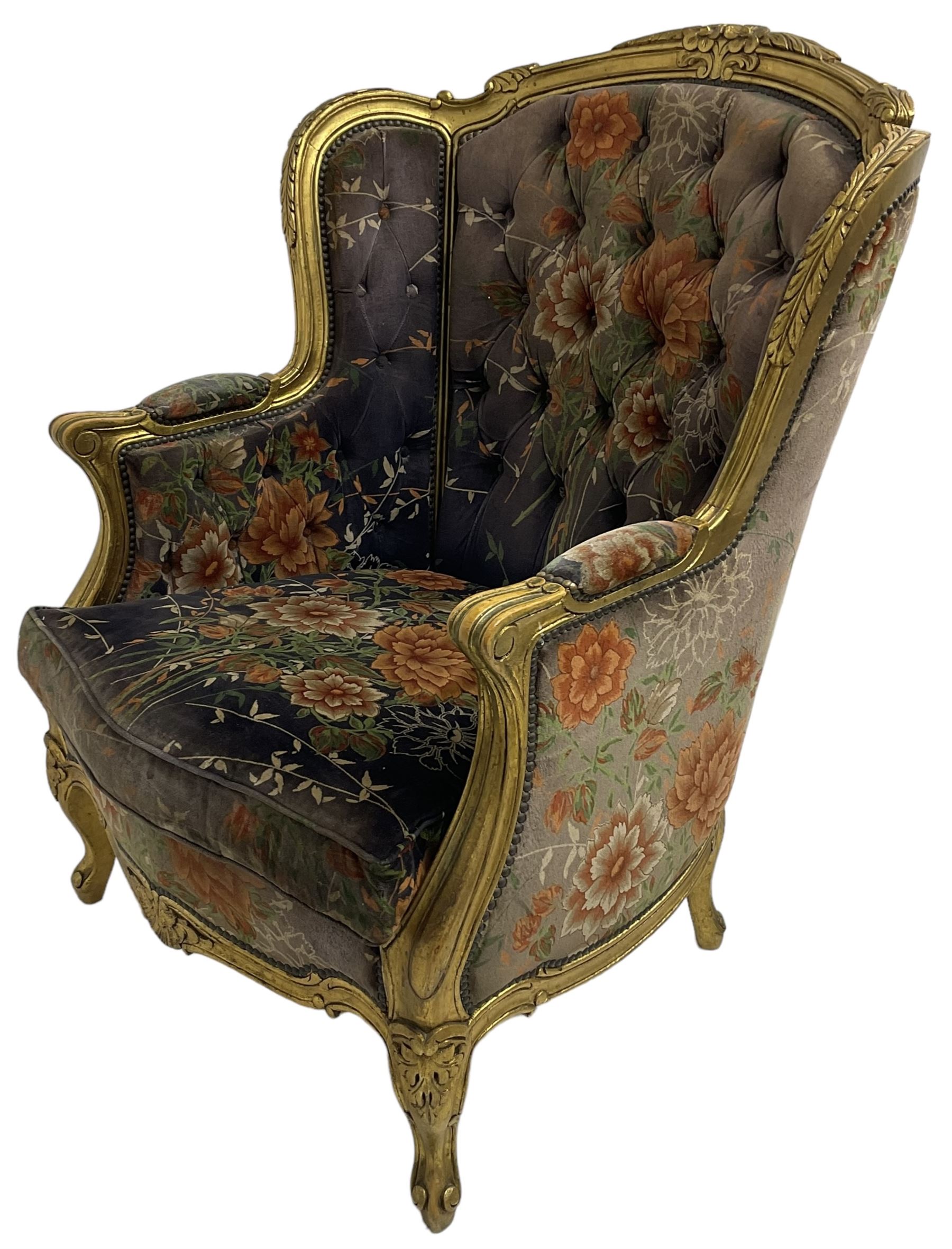 Pair of Louis XVI design gilt framed wingback armchairs, cresting rail carved and moulded with foliate decoration and C-scrolls, upholstered in buttoned floral patterned mauve velvet with loose seat cushion, scallop carved apron over cabriole supports