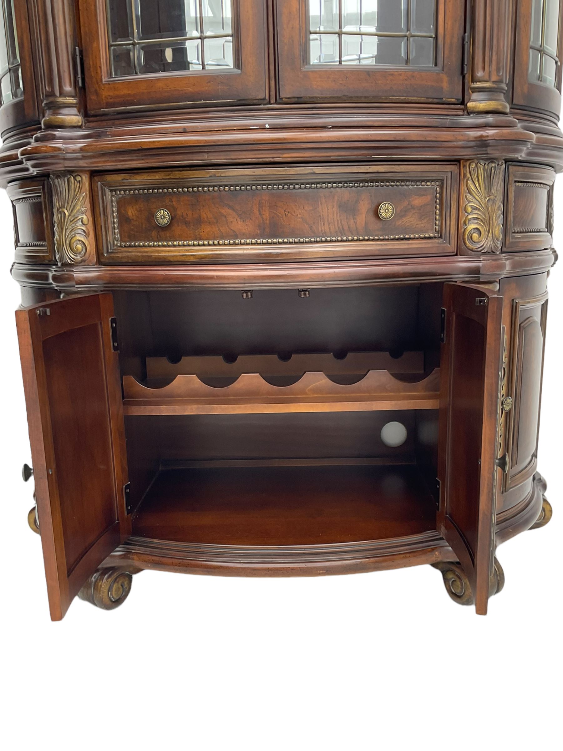  Kevin Charles American walnut demi lune display cabinet with illuminated interior, projecting cornice, two bevel edge glazed doors enclosing two adjustable shelves above single drawer and four cupboard doors with wine rack, carved scrolling supports