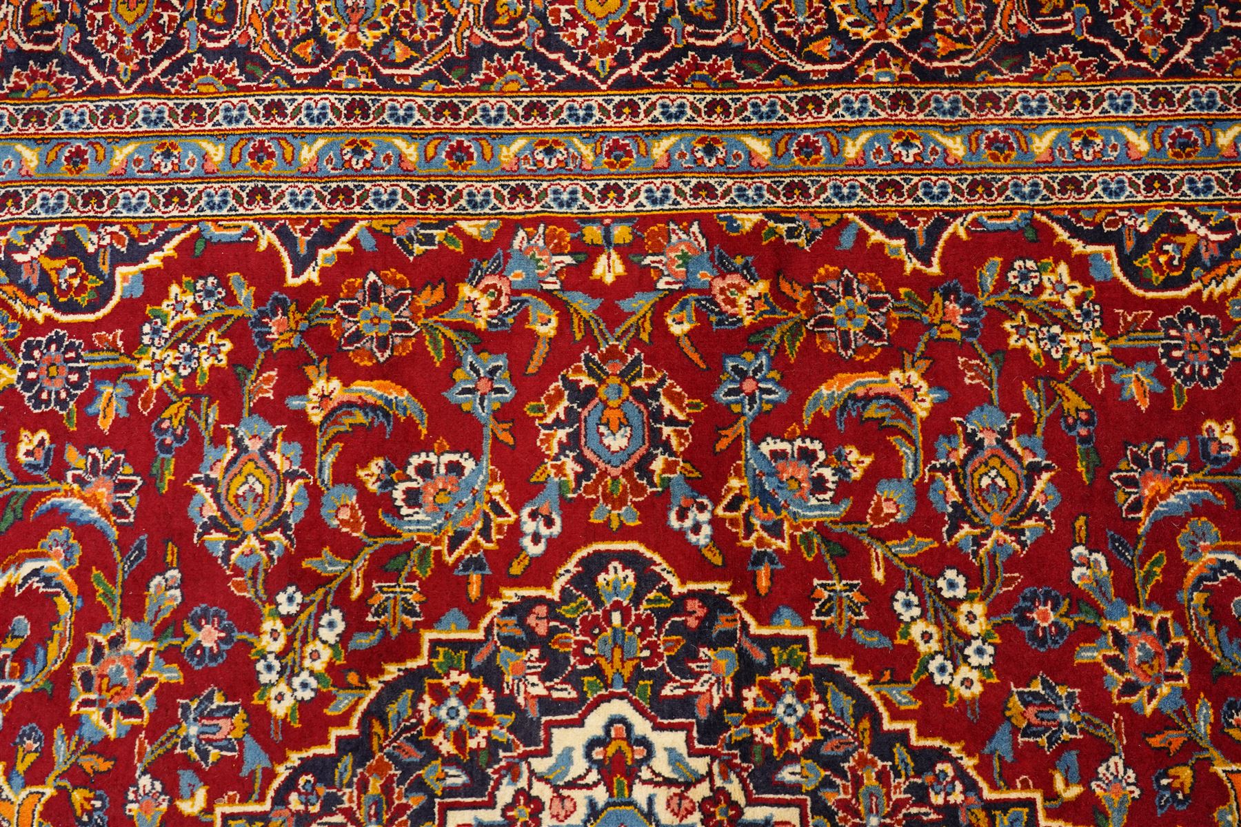 Central Persian Kashan hand-knotted carpet, crimson field with central cusped medallion in ivory, pale blue and indigo, issuing a network of scrolling arabesques, palmettes, and floral sprays, with conforming spandrels, main border of scrolling vinery and stylized palmettes, framed by multiple guard borders, with cotton fringe ends 