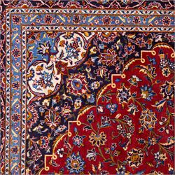 Central Persian Kashan hand-knotted carpet, rich crimson red field with a central cusped medallion with finely drawn floral sprays surrounded by dense scrolling vine work and palmettes, framed by a broad indigo ground border of flowering arabesques flanked by multiple guard stripes, in a palette of red, blue, ivory, sage green, and soft ochre tones, with cotton fringe ends    