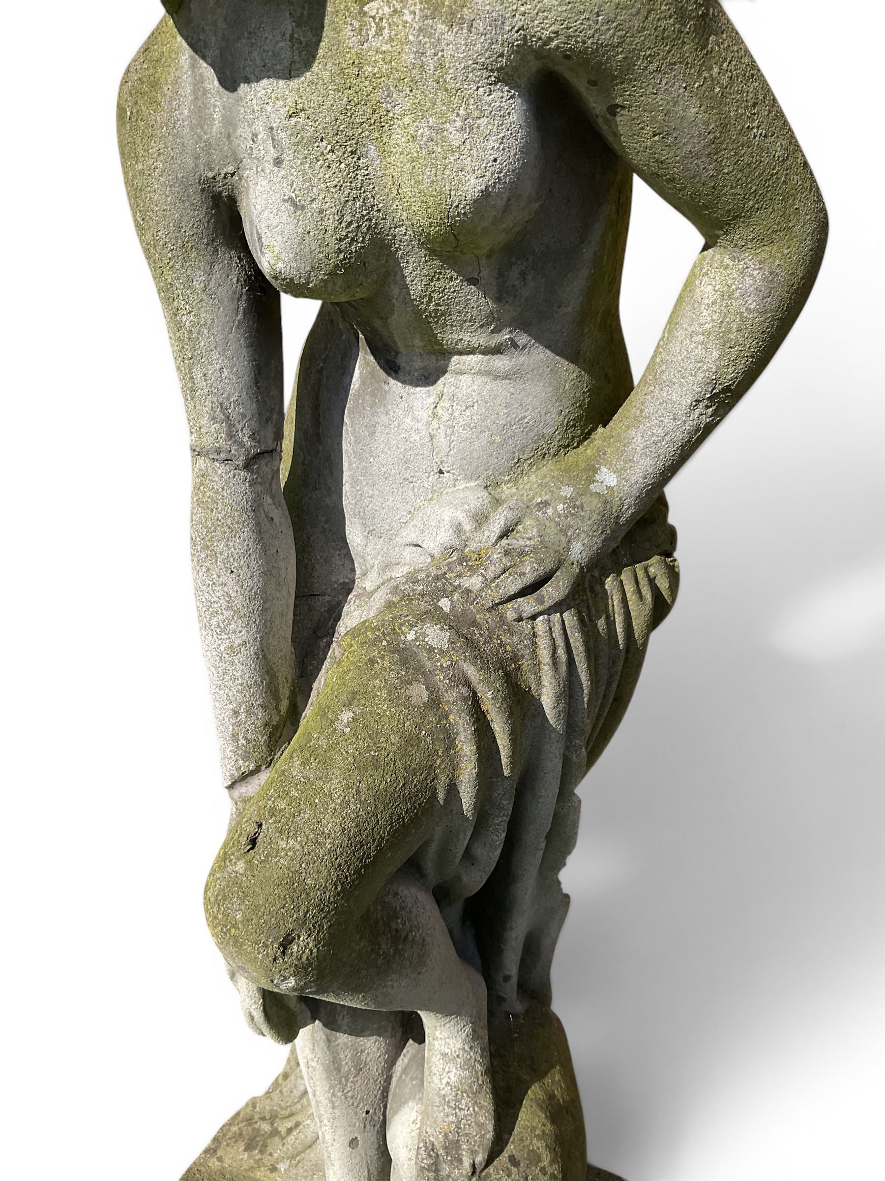 Cast stone garden figure of a classically draped nude female inspired by Aphrodite statues, with stylised up-do arranging her chitin on a circular base, the rectangular pedestal with egg-and-dart moulding over a floral wreath, in weathered finish