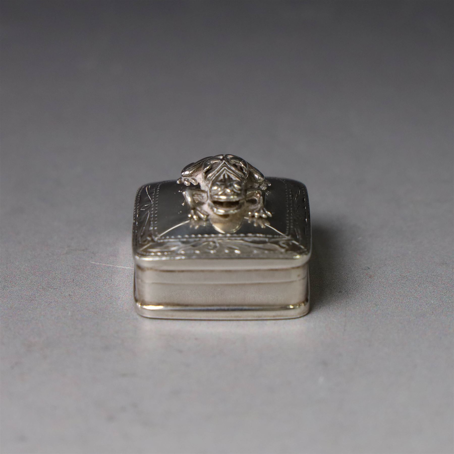 Modern silver pill box, of square form, the hinged cover surmounted with figure of a frog within a engraved scroll edge, hallmarked hallmarked Martin Gerald & Sid Adler, London 2000, W3cm