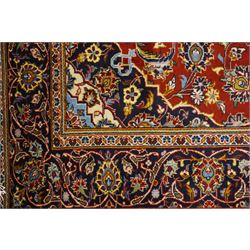 Central Persian Kashan hand-knotted rug, madder red field with central articulated cusped medallion in ivory and sky blue, issuing network of scrolling arabesques palmettes and floral sprays with conforming spandrels, indigo main border of scrolling vinery and stylized palmettes, framed by multiple guard borders, with cotton fringe ends  
