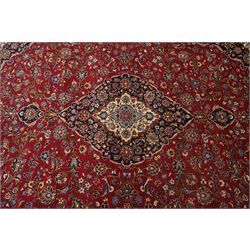 Central Persian Kashan hand-knotted carpet, crimson ground field, with central cusped lozenge medallion, enclosing a palmette and a floral rosette arrangement, surrounding field densely populated with scrolling arabesques, flowering vinery and stylized boteh motifs in tones of indigo, pale blue, ivory and ochre and spandrels of confirming design, flanked by multiple guard stripes, with cotton fringe ends 