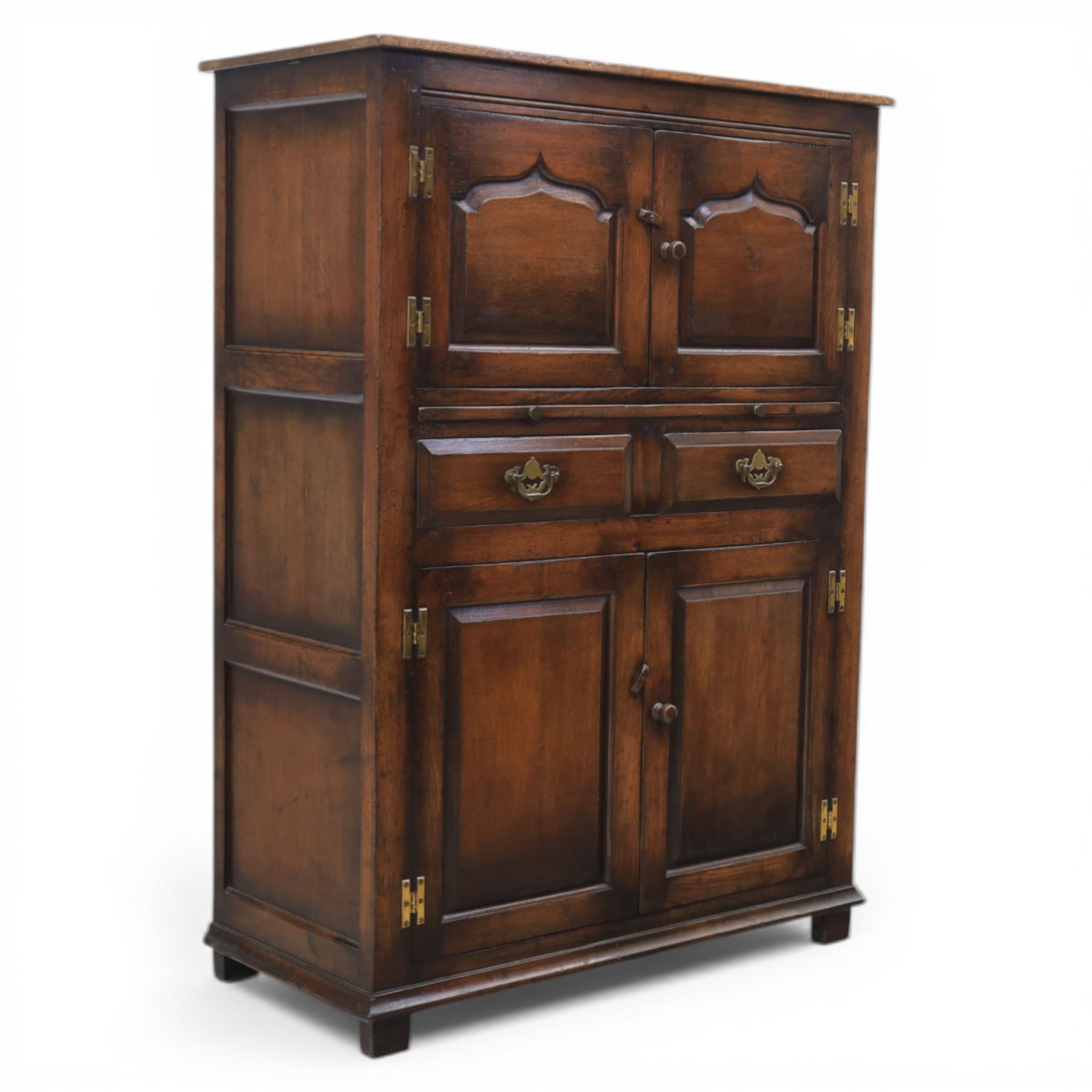  Late 20th century oak four door cupboard in the manner of Titchmarsh and Goodwin, rectangular plank top above two pairs of fielded panel doors with shaped raised panels, central arrangement of a brushing slide above two short drawers with brass swan neck handles and pierced backplates, enclosed by further pair of panelled cupboard doors, fitted with brass butt hinges and turned wooden knobs, raised on plinth base