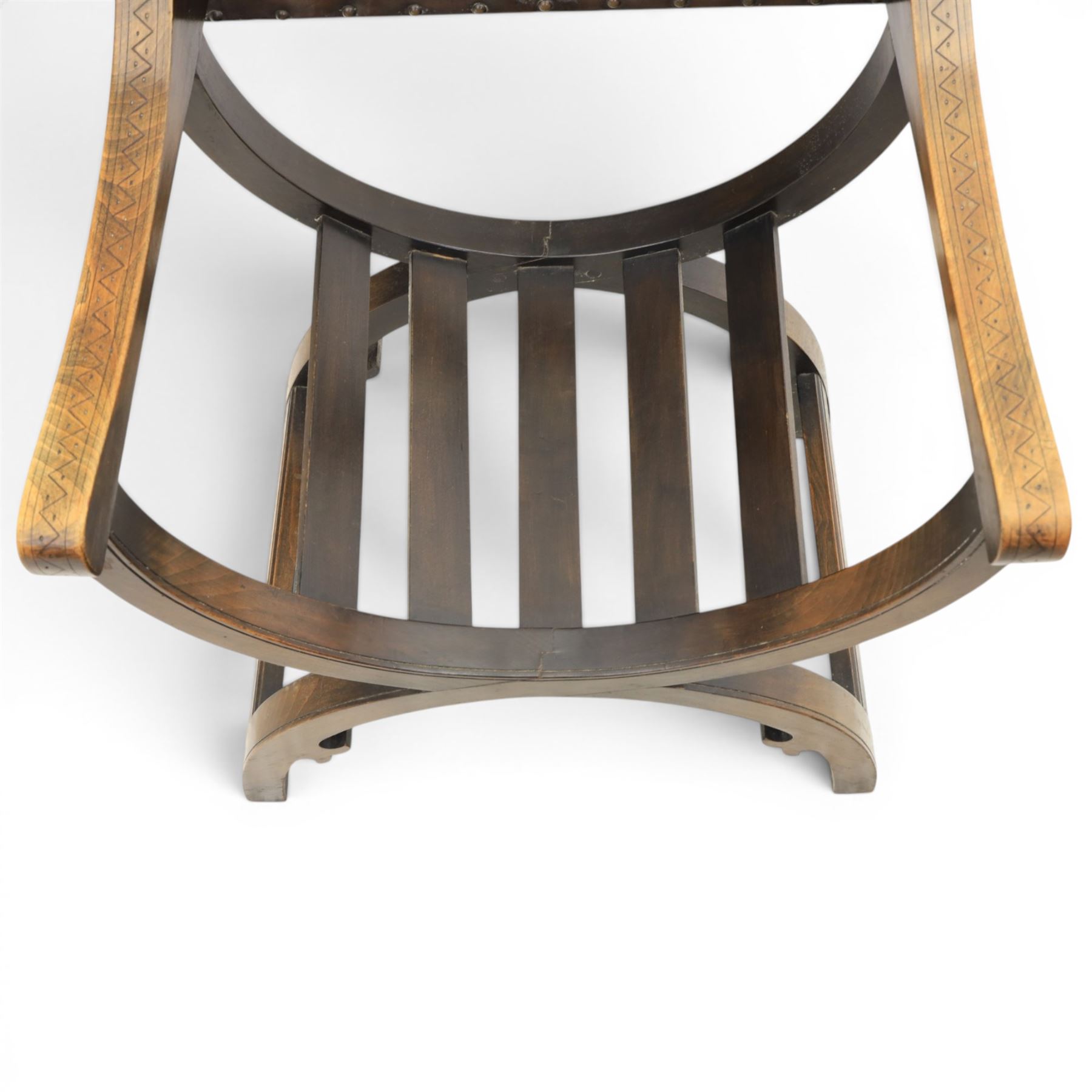 Early 20th century mahogany Savonarola chair, arched leather back with brass studwork, curved open arms with incised geometric decoration, loose patchwork velvet cushion seat, X-frame supports united by circular side rails, on scrolled feet