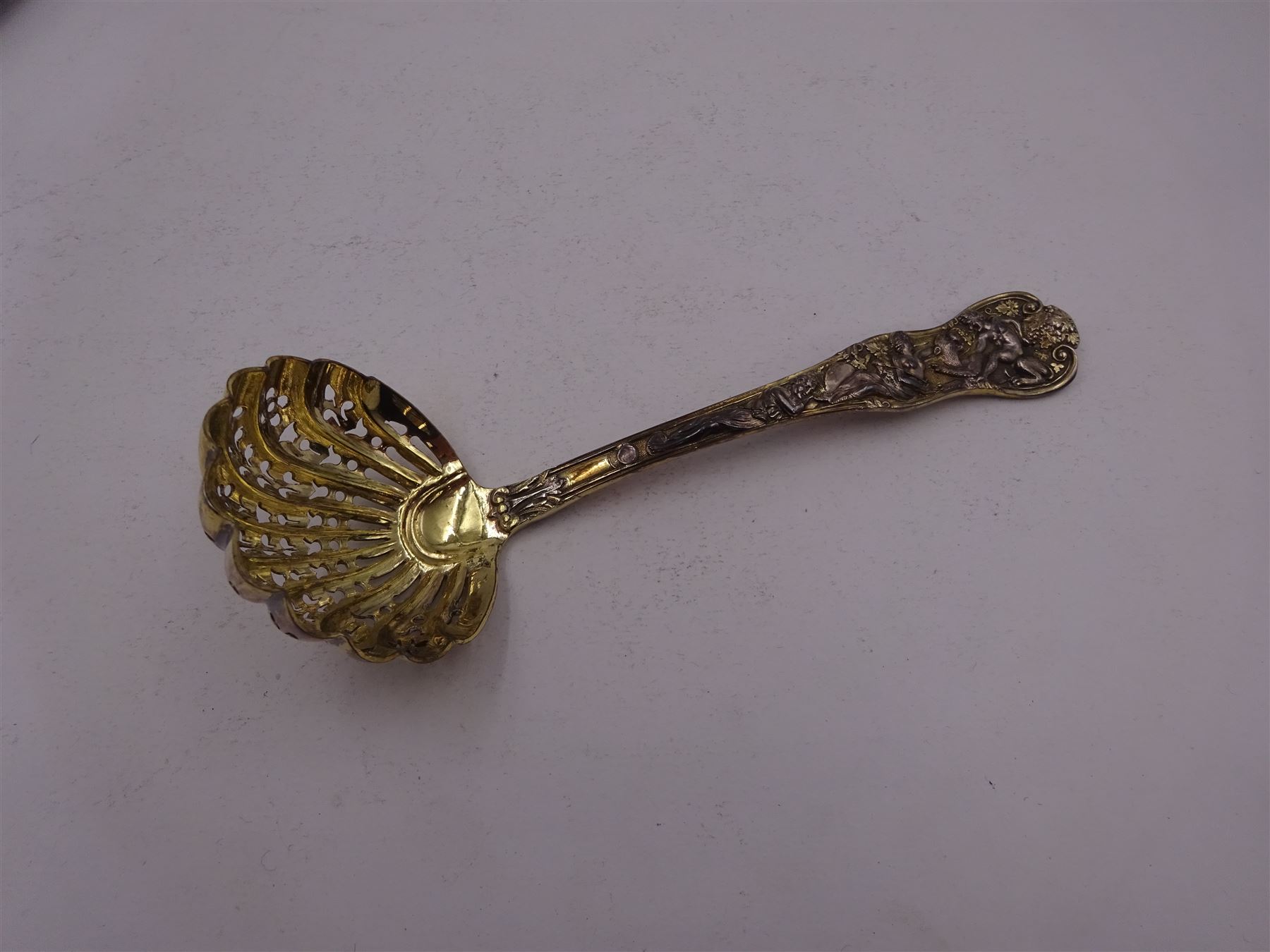 Victorian silver-gilt sugar sifting spoon, with gilded pierced shell shaped bowl, the handle decorated in relief with classical greek figures amongst fruiting vines, hallmarked Henry John Lias & James Wakely, London 1874,  L16cm, contained within tooled leather silk and velvet lined fitted case