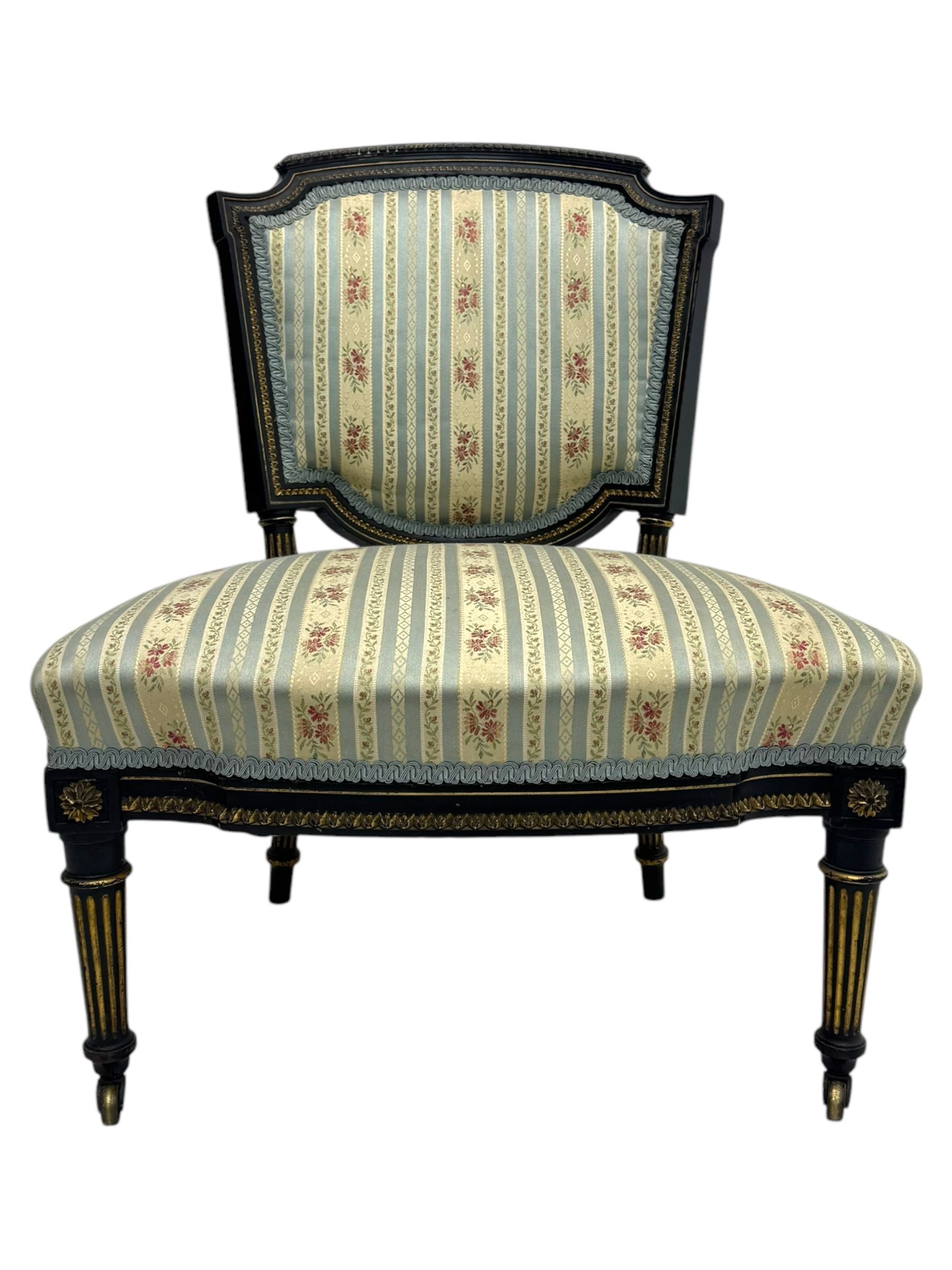 19th century French ebonised low chair, upholstered in floral striped fabric within a moulded frame with gilt line and foliate decoration, the seat of serpentine form, raised on turned fluted legs with toupie feet