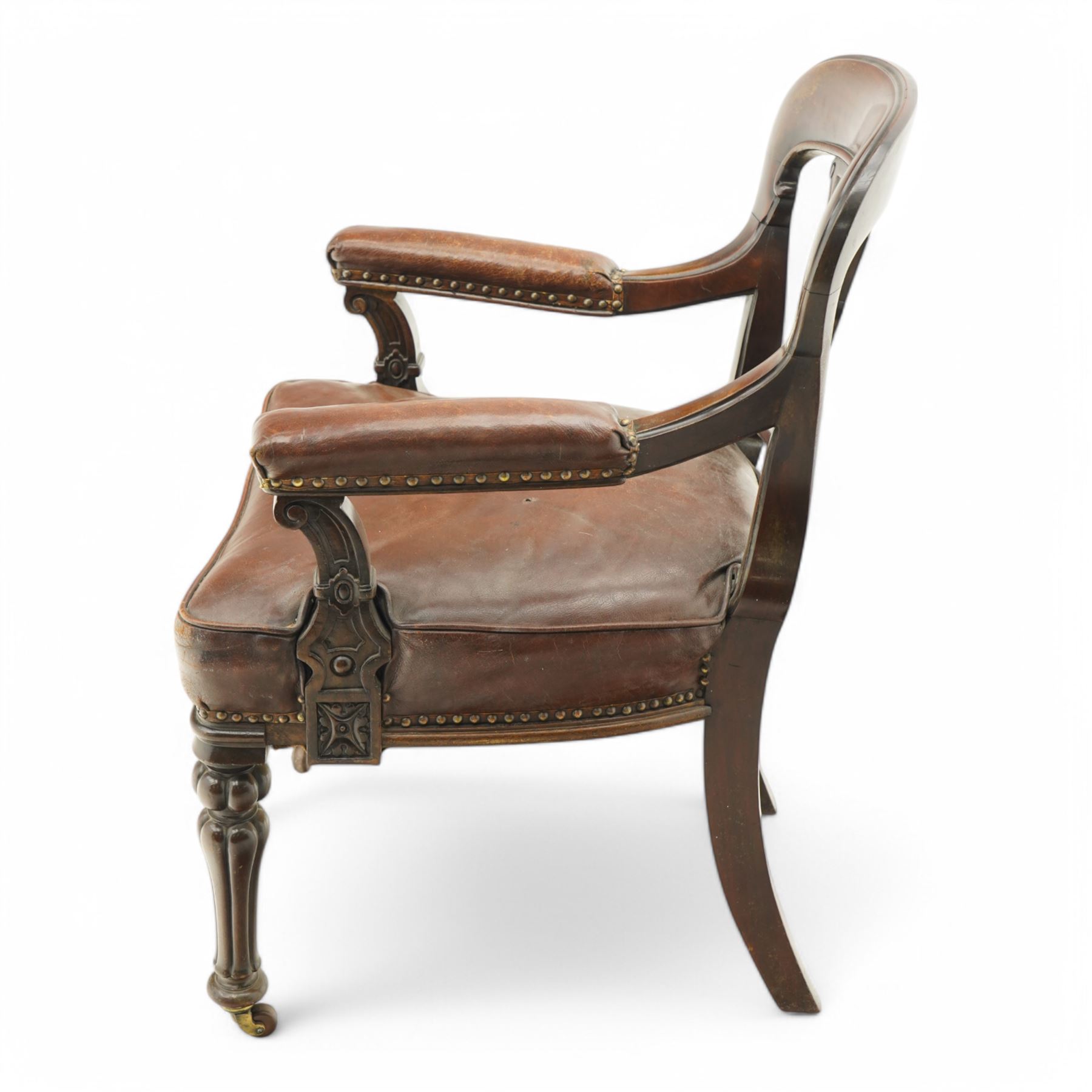 Victorian mahogany library elbow chair, arched cresting rail over shaped and pierced splat carved with cartouche, upholstered arms and seat in brown leather with studded bands, turned and lobe carved front supports on brass and ceramic castors 