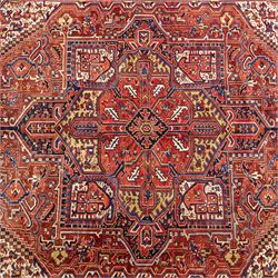 North West Persian Heriz hand-knotted carpet, madder red field, with a large angular stepped central medallion with serrated outlines, flanked by bold stylized palmettes, hooked leaves and geometric devices, spandrels similarly decorated within a wide geometric main border featuring angular floral motifs, enclosed by multiple guard borders in madder red, indigo blue, ivory, olive green and soft ochre, with cotton fringe ends 