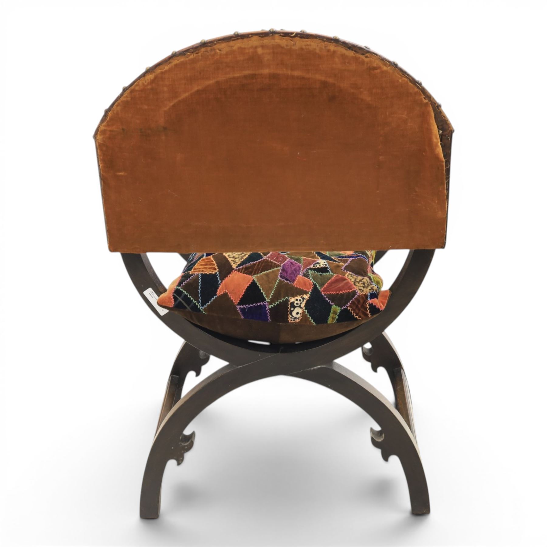 Early 20th century mahogany Savonarola chair, arched leather back with brass studwork, curved open arms with incised geometric decoration, loose patchwork velvet cushion seat, X-frame supports united by circular side rails, on scrolled feet