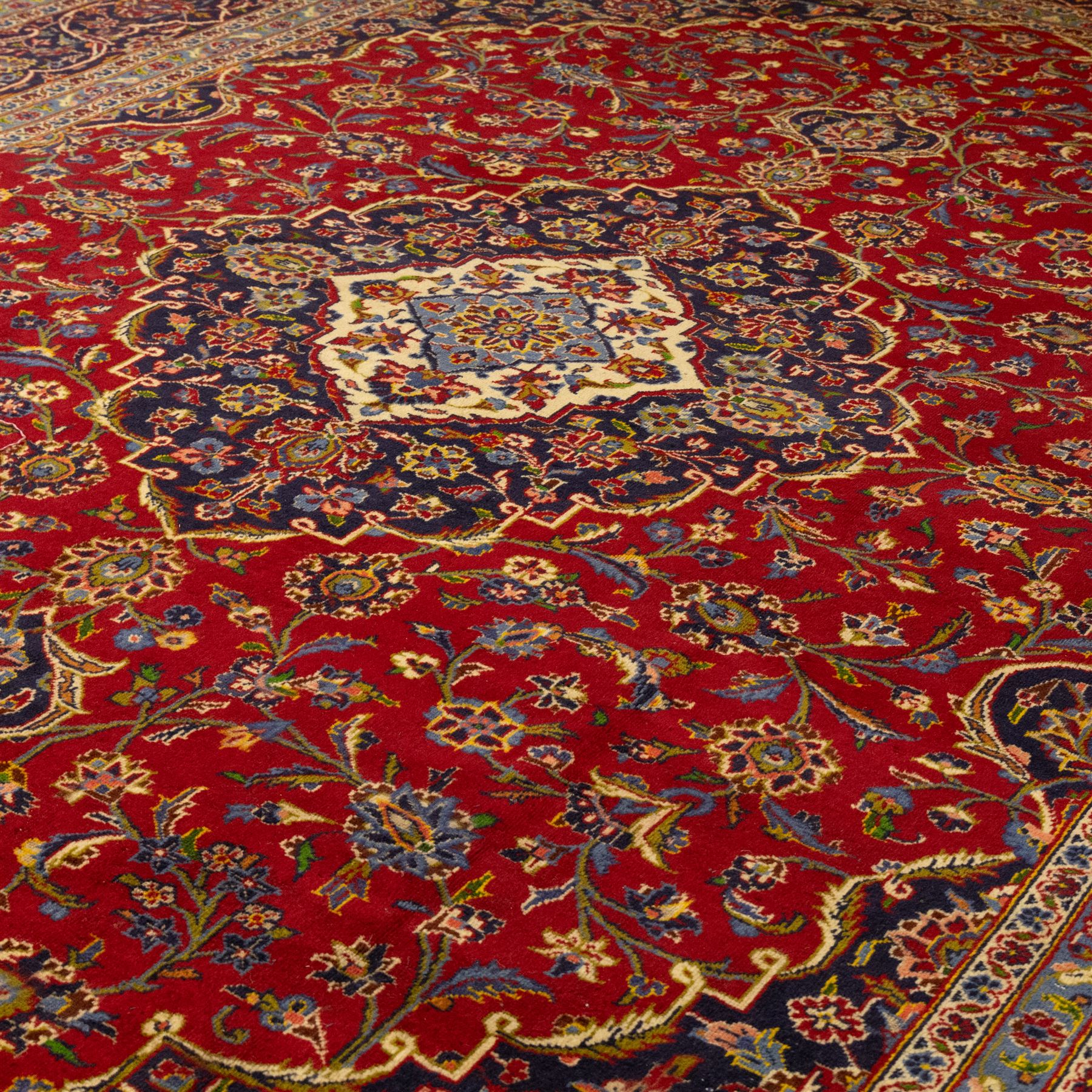 Central Persian Kashan hand-knotted carpet, crimson ground, with a central shaped palmette medallion with pendants, surrounded by scrolling foliate and floral motifs incorporating rosettes, palmettes, and vine work, enclosed by a dark navy with repeating border with repeating palmettes and meandering vines, flanked by floral guard stripes, with cotton fringes 