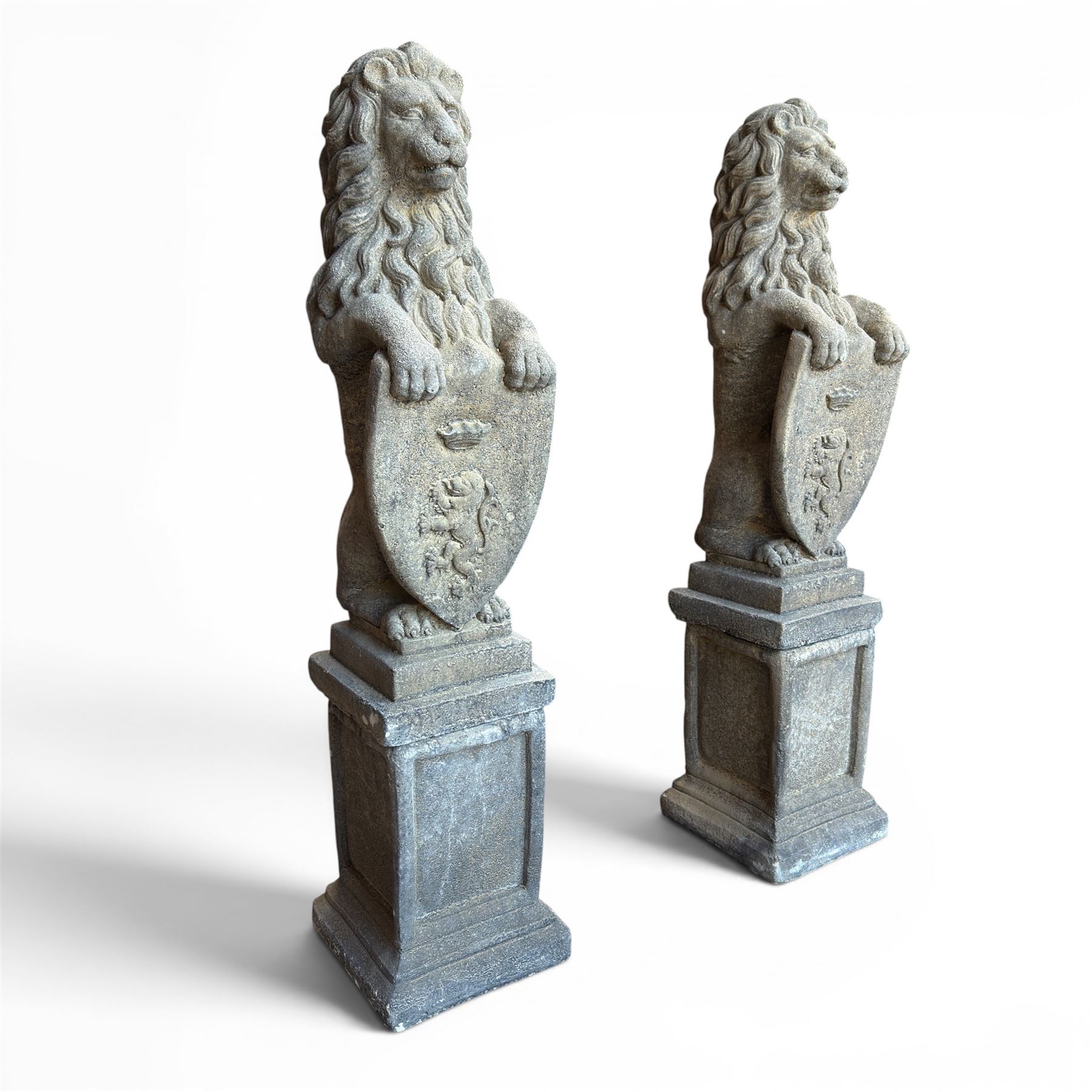 Pair of cast stone Heraldic lions, modelling in upright standing poses with flowing mane, each clasping an Armorial shield moulded with a crowned lion in their forepaws, raised on rectangular pedestals