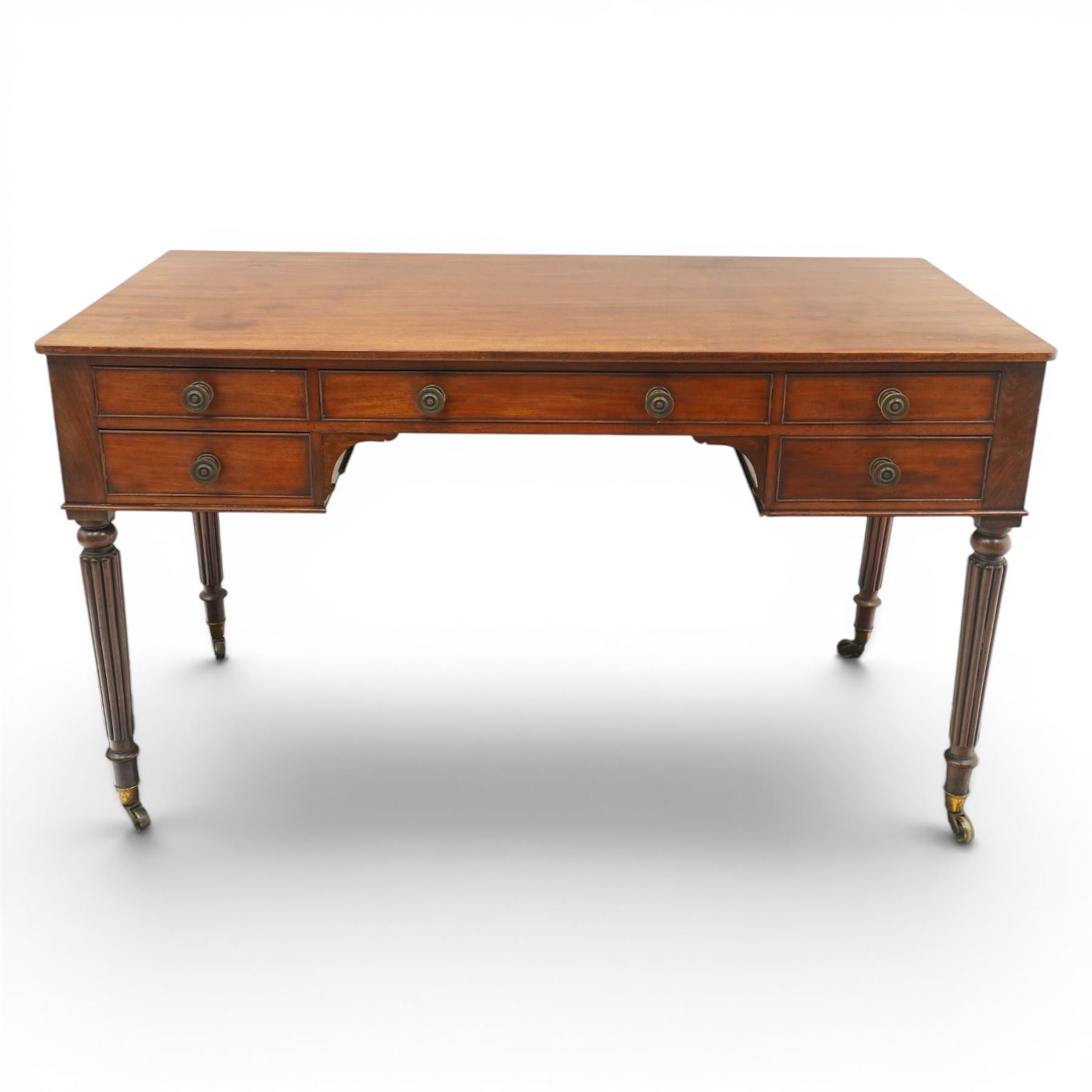 In the manner of Gillows - Regency period mahogany kneehole writing table, rectangular top over central cock-beaded frieze drawer with shaped spandrels, flanked by two graduating drawers to either side, raised on turned and fluted supports terminating to brass cups and castors