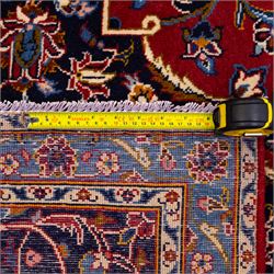 Central Persian Kashan hand-knotted carpet, rich crimson red field with a central cusped medallion with finely drawn floral sprays surrounded by dense scrolling vine work and palmettes, framed by a broad indigo ground border of flowering arabesques flanked by multiple guard stripes, in a palette of red, blue, ivory, sage green, and soft ochre tones, with cotton fringe ends    
