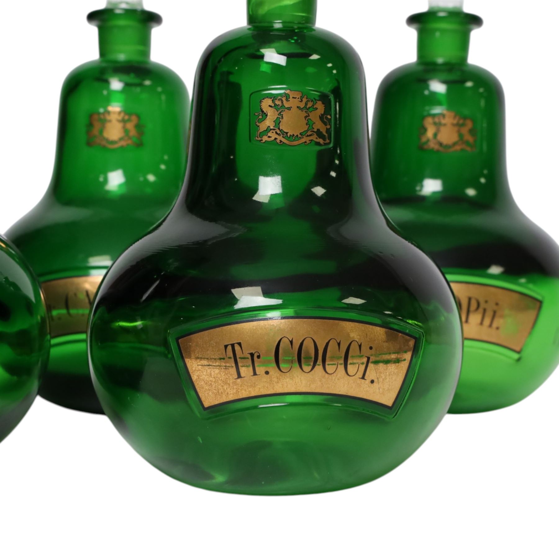 Set of four 20th century Royal Pharmaceutical Society green glass bottles, of bellied form with clear cut glass spire stopper, each body bearing gilt label in Latin: Tr.OPii., Tr.COCCi., Tr.CANNAB., and Tr.LOBEL., and gilt Royal Pharmaceutical Society crest to neck, H25cm

Provenance: From the collection of the family of Peter Green, a practising pharmacist whose career spanned the mid to late 20th century. During his professional life he managed pharmacies in Yeovil, Somerset, and later Selles Chemist in Filey, North Yorkshire, before purchasing and running his own business, Peter Green Pharmacy, located in the Market Place, Malton. The bottles were displayed as part of the traditional shop fittings in both the Filey and Malton pharmacies and were retained as part of his collection.