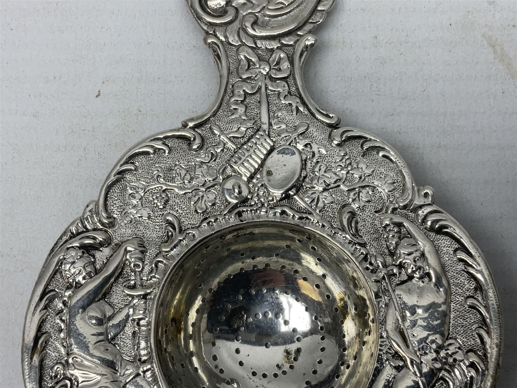 Continental white metal strainer, of shaped form embossed with classical figures, putto, musical trophies and fruiting vines, indistinctly marked, L12.5cm