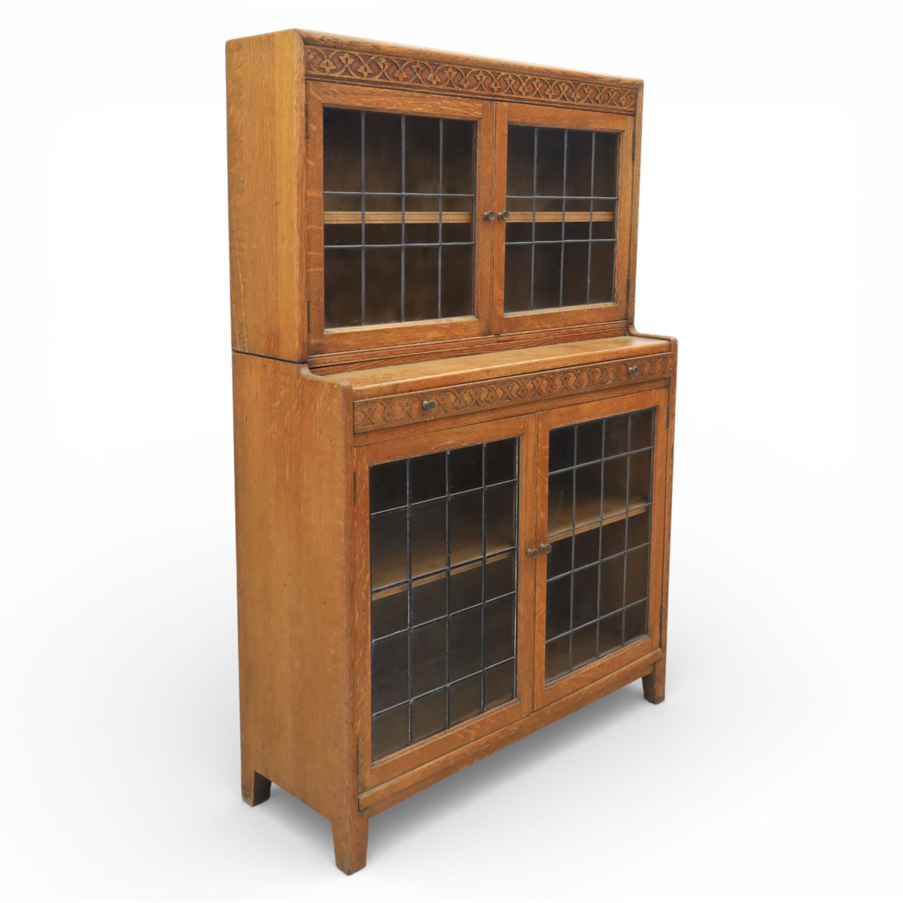 Early 20th-century Arts and Crafts glazed oak bookcase, upper section fitted with a pair of leaded glazed doors enclosing a fixed shelf above a projecting frieze drawer interior with baize-lined writing surface flanked by two compartments, carved with interlaced decoration, lower section fitted with a further pair of leaded glazed doors enclosing a single shelf, raised on short stile feet 