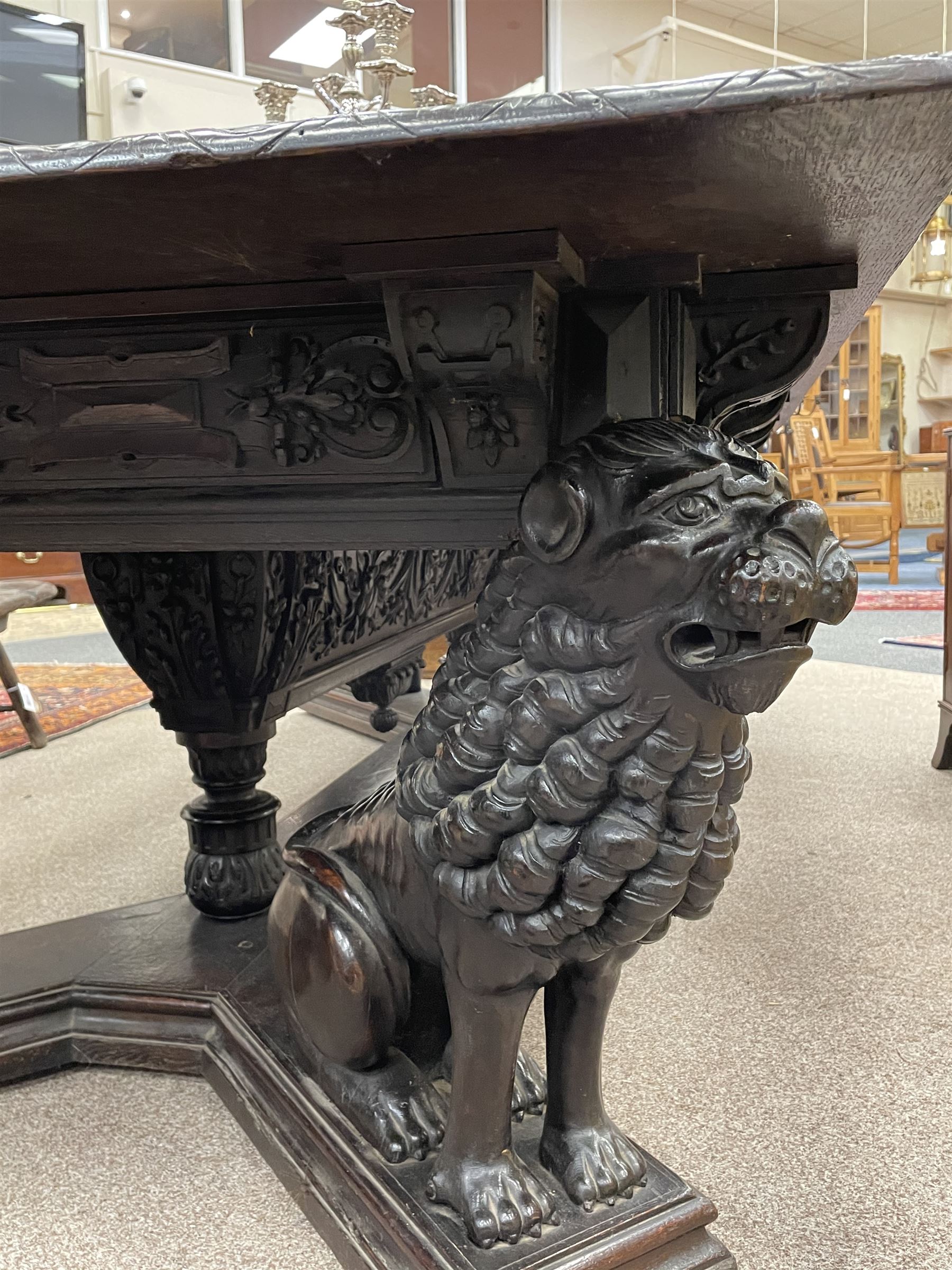 Large 19th century carved oak library table, the rectangular top with lunette carved edge over cartouche panelled and carved  scroll frieze, the central tapering rectangular support relief carved with masks and garlands, on four carved seated lion supports at each corner, on flat angular stretcher with recessed castors stamped ‘Bartholomew & Fletcher, London’