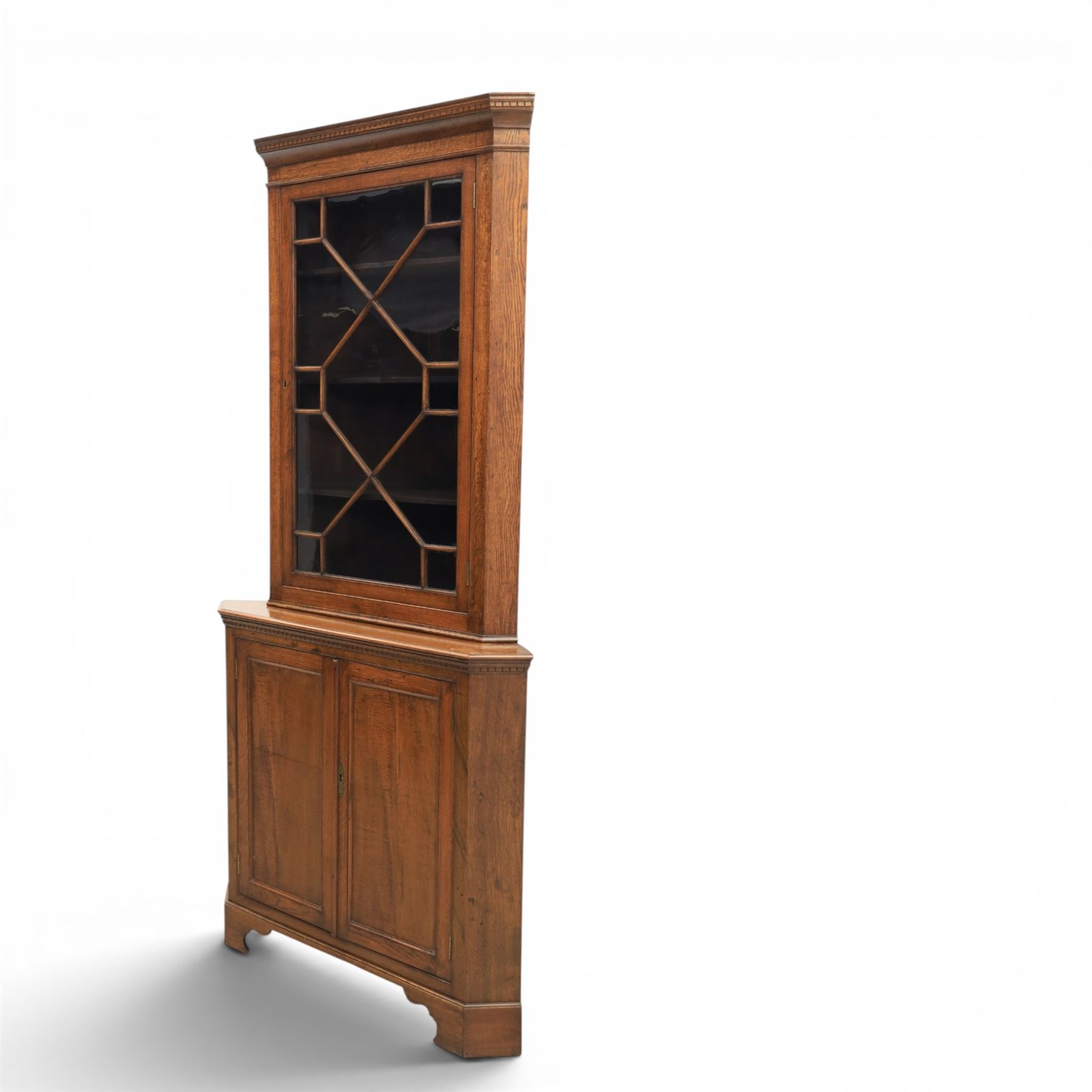19th century oak floor-standing corner display cabinet, dentil cornice above astragal glass, shaped fixed shelves, the lower section with twin cupboard doors, on shaped bracket feet